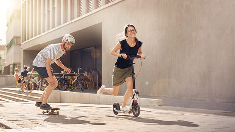 Mann auf Skateboard und Frau auf Tretroller