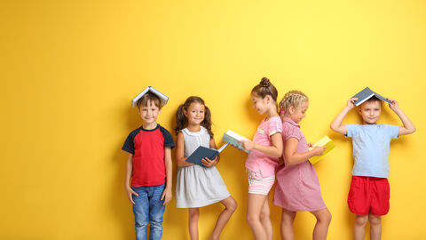 Kinder stehen vor gelber Wand und lesen in Büchern oder haben Bücher auf dem Kopf