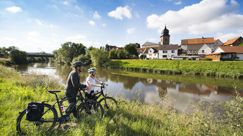 Ein Mann und eine Frau stehen mit Elektrofahrrädern an der Fulda und schauen auf ein Dorf