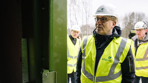 Energieminister Al-Wazir in gelber Jacke und mit Helm bei der Baustellenbesichtigung vor einem Windrad.