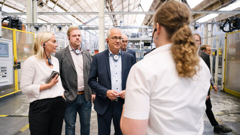 Im Volkswagen Werk stehen Wirtschaftsminister Al-Wazir und weitere Personen mit Kopfhörern in einer Halle. Eine weitere Person ist ihnen zugewandt und erklärt.