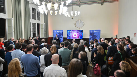 Empfang der Kreativwirtschaft  Mehrere Personen stehen vor einer Empore und hören einem Vortrag zu