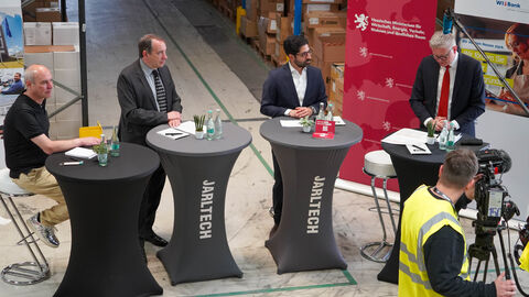 Vier Personen stehen in einer Lagerhalle an Stehtischen