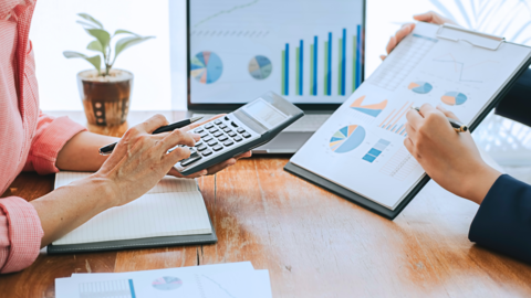 Wirtschaft Eine Person rechnet mit einem Taschenrechner eine andere Person hält ein Klemmbrett mit Diagrammen