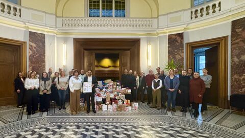 Mehrere Personen stehen in einem großen Raum. In der Mitte liegen ganz viele verpackte Geschenke