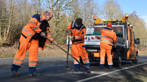Drei Straßenarbeiter in orangener Kleidung reparieren ein Schlagloch